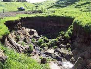 Grassy land surrounds a large, deep gully. The top soil has been eroded by water and the land has caved in. Water, soil and grass are in the gully. 